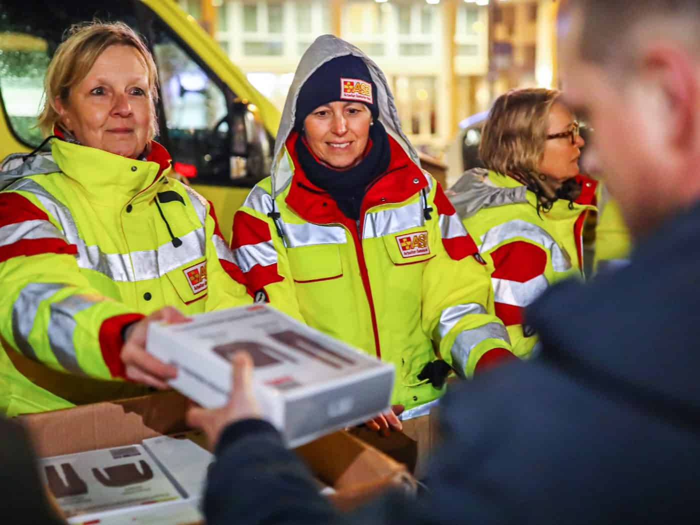 Zwei ASB-Helferinnen in gelb-roter Einsatzkleidung händigen im Freien ein Paket mit Unterwäsche/Gegenständen aus einem Karton an eine Person aus. Eine weitere Helferin steht im Hintergrund vor einem gelben ASB-Fahrzeug.