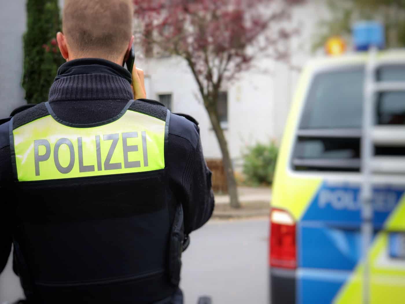 Person in schwarzer Uniform mit neon-gelber „POLIZEI"-Weste hält ein Funkgerät in der Hand, im Hintergrund ein Streifenwagen mit blauer und gelber Markierung