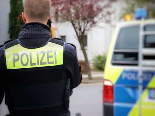 Person in schwarzer Uniform mit neon-gelber „POLIZEI"-Weste hält ein Funkgerät in der Hand, im Hintergrund ein Streifenwagen mit blauer und gelber Markierung