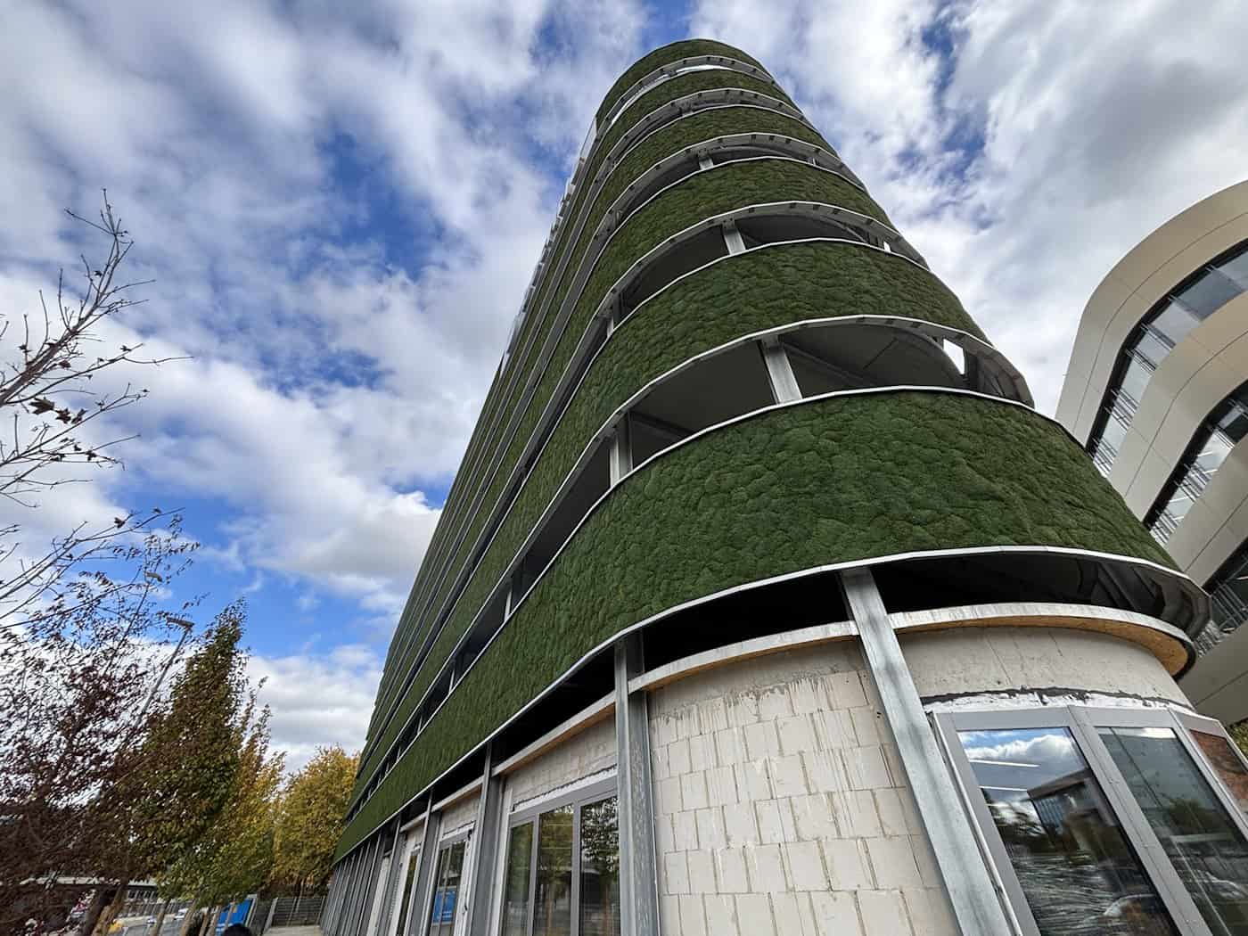 Steile Froschperspektive auf ein modernes, mehrstöckiges Parkhaus mit sattgrünen, moosbedeckten Fassadenbändern. Die geschwungene Gebäudeform ragt dynamisch vor einem leicht bewölkten blauen Himmel auf.