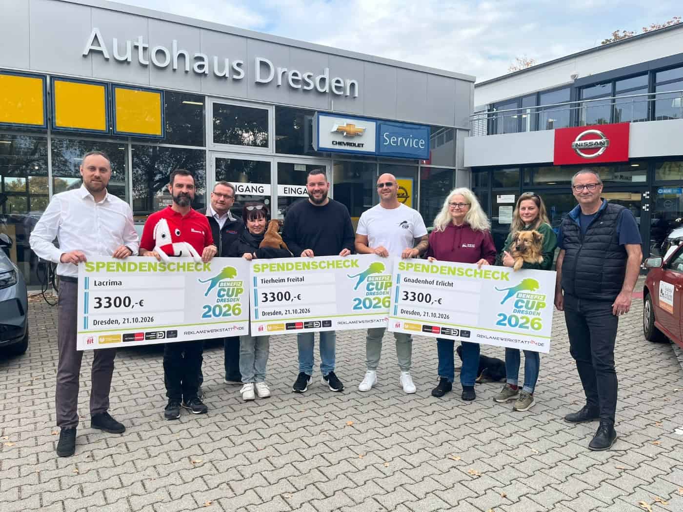 Neun Personen stehen mit drei großen Spendenschecks über jeweils 3.300 Euro und zwei Hunden vor dem Autohaus Dresden.