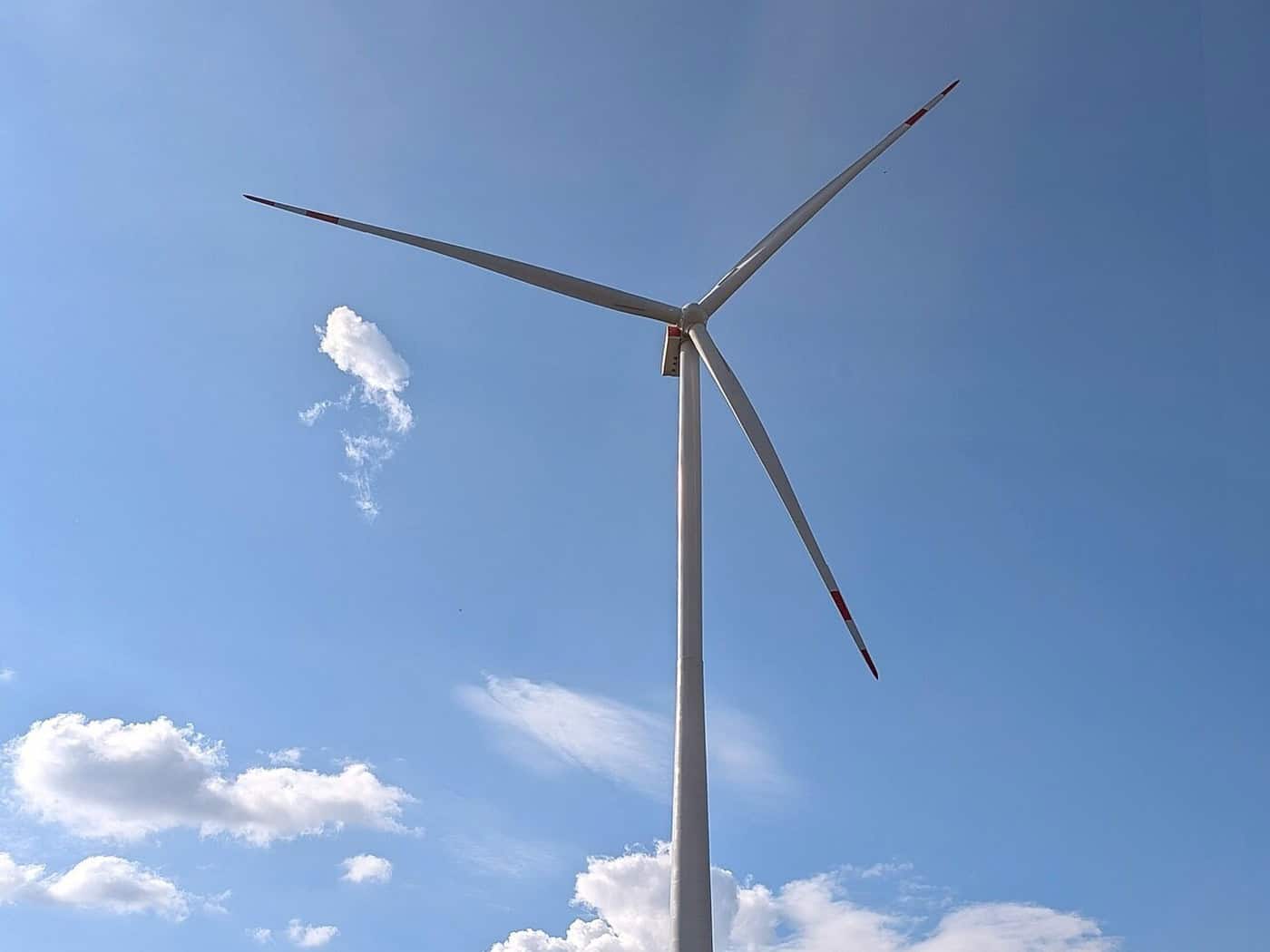 Weiße Windenergieanlage mit roten Markierungen an Turm und Rotorblättern vor blauem Himmel, unten Waldrand
