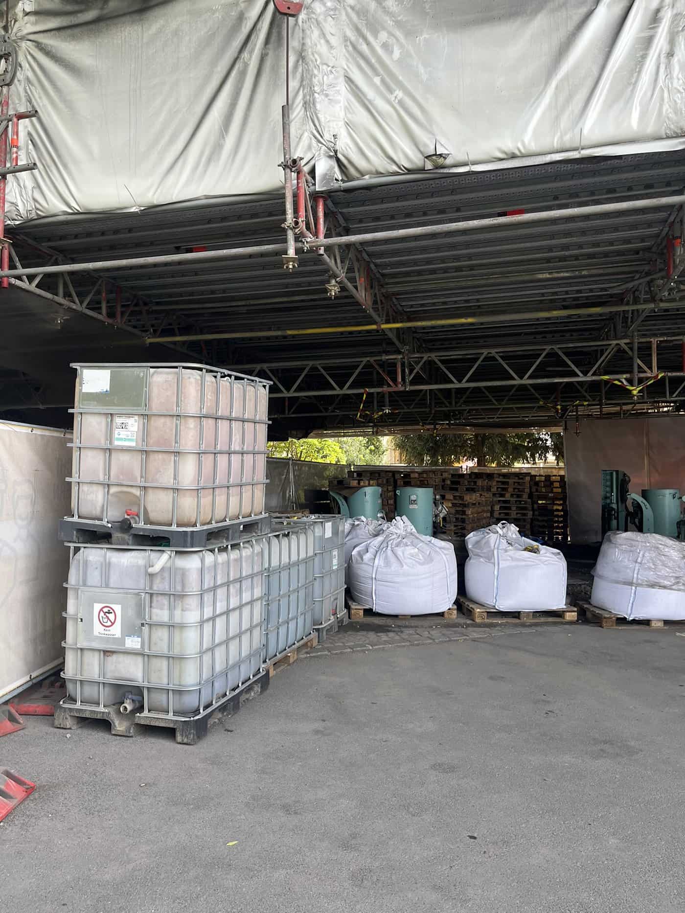 Blick unter ein Baugerüst am Blauen Wunder: Stapel von Paletten, große weiße Big Bags mit Baumaterial sowie zwei aufeinandergestapelte Plastik-Wassertanks stehen auf dem gepflasterten Boden.