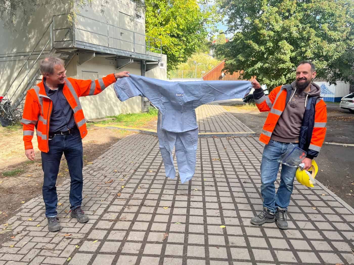 Bauingenieur Christoph Nollau und Projektleiter Robert Bauch halten gemeinsam einen hellblauen Ganzkörper-Schutzanzug vor einem Baucontainer hoch. Beide tragen Warnjacken und lächeln in die Kamera.
