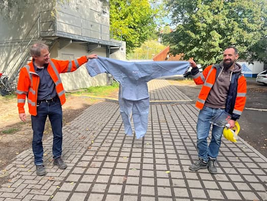 Bauingenieur Christoph Nollau und Projektleiter Robert Bauch halten gemeinsam einen hellblauen Ganzkörper-Schutzanzug vor einem Baucontainer hoch. Beide tragen Warnjacken und lächeln in die Kamera.