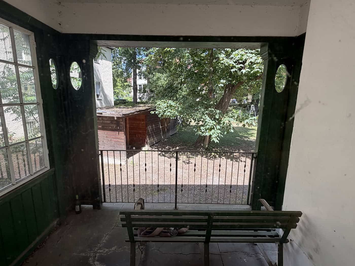 Blick aus einer überdachten Loggia oder von einem Balkon durch ein Geländer auf ein weitläufiges Gartengrundstück mit alten Laubbäumen und einer kleinen Holzhütte im Hintergrund.