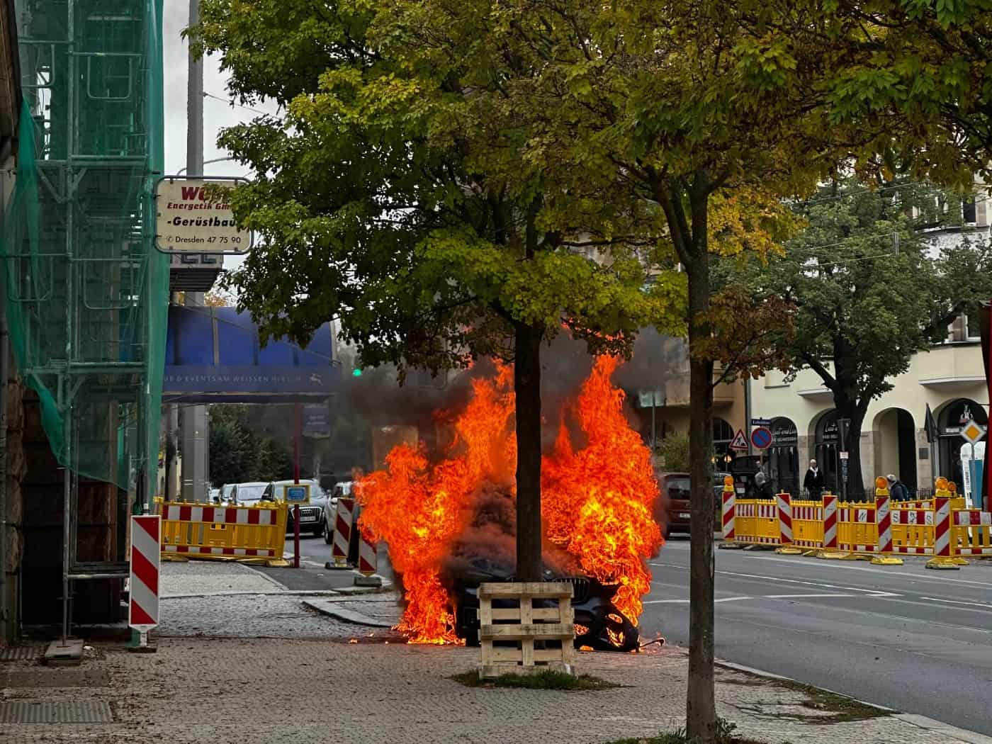 Ein auf der Straße stehendes Auto schlägt auf dem Weißen Hirsch in Dresden meterhoch in hellen Flammen und dickem dunklem Rauch; im Vordergrund stehen Bäume, ein gepflasterter Gehweg mit Gerüst und rot-weiße Absperrgitter.