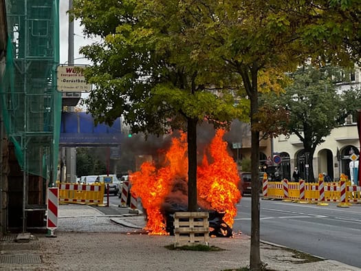 Ein auf der Straße stehendes Auto schlägt auf dem Weißen Hirsch in Dresden meterhoch in hellen Flammen und dickem dunklem Rauch; im Vordergrund stehen Bäume, ein gepflasterter Gehweg mit Gerüst und rot-weiße Absperrgitter.