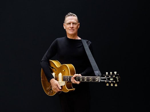 Studio-Porträt des Musikers Bryan Adams vor schlichtem, dunklem Hintergrund. Er trägt ein schwarzes Langarmshirt, blickt direkt in die Kamera und hält eine goldfarbene Gibson-Gitarre vor seinem Körper.