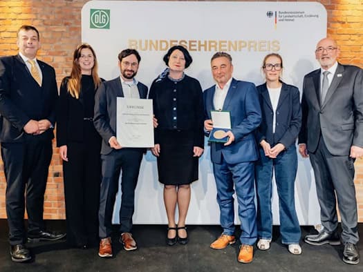 Sieben Personen stehen vor einer Wand mit der Aufschrift „Bundesehrenpreis“ und den Logos von DLG und Bundeslandwirtschaftsministerium; ein Mann hält eine Urkunde, ein anderer eine Medaille im Etui.