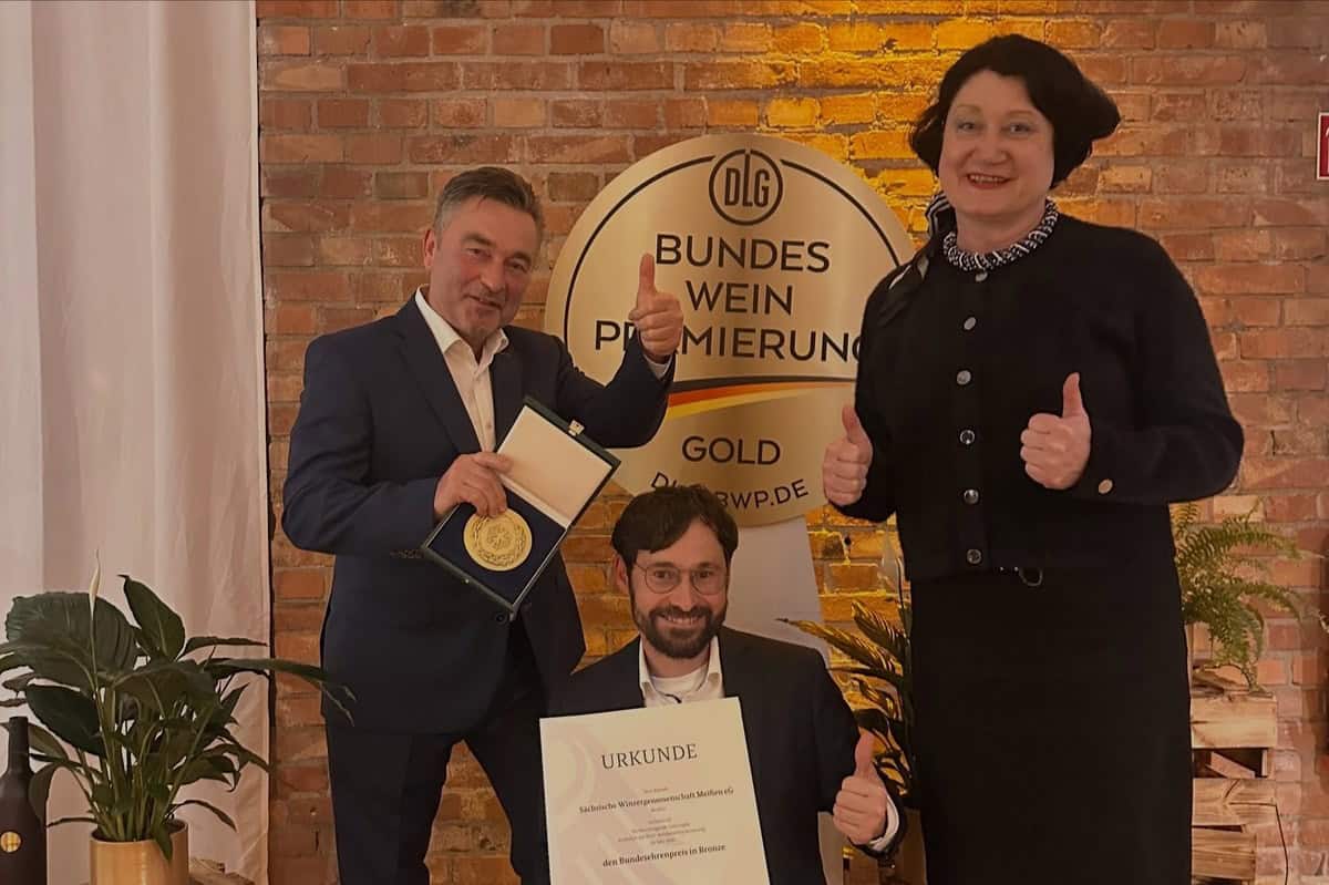 Bundesehrenpreis in Bronze für die Winzergenossenschaft Meißen Ein Mann mit Medaille im Etui, ein knieender Mann mit Urkunde und eine Frau zeigen vor einem Schild der DLG-Bundesweinprämierung die Daumen nach oben.