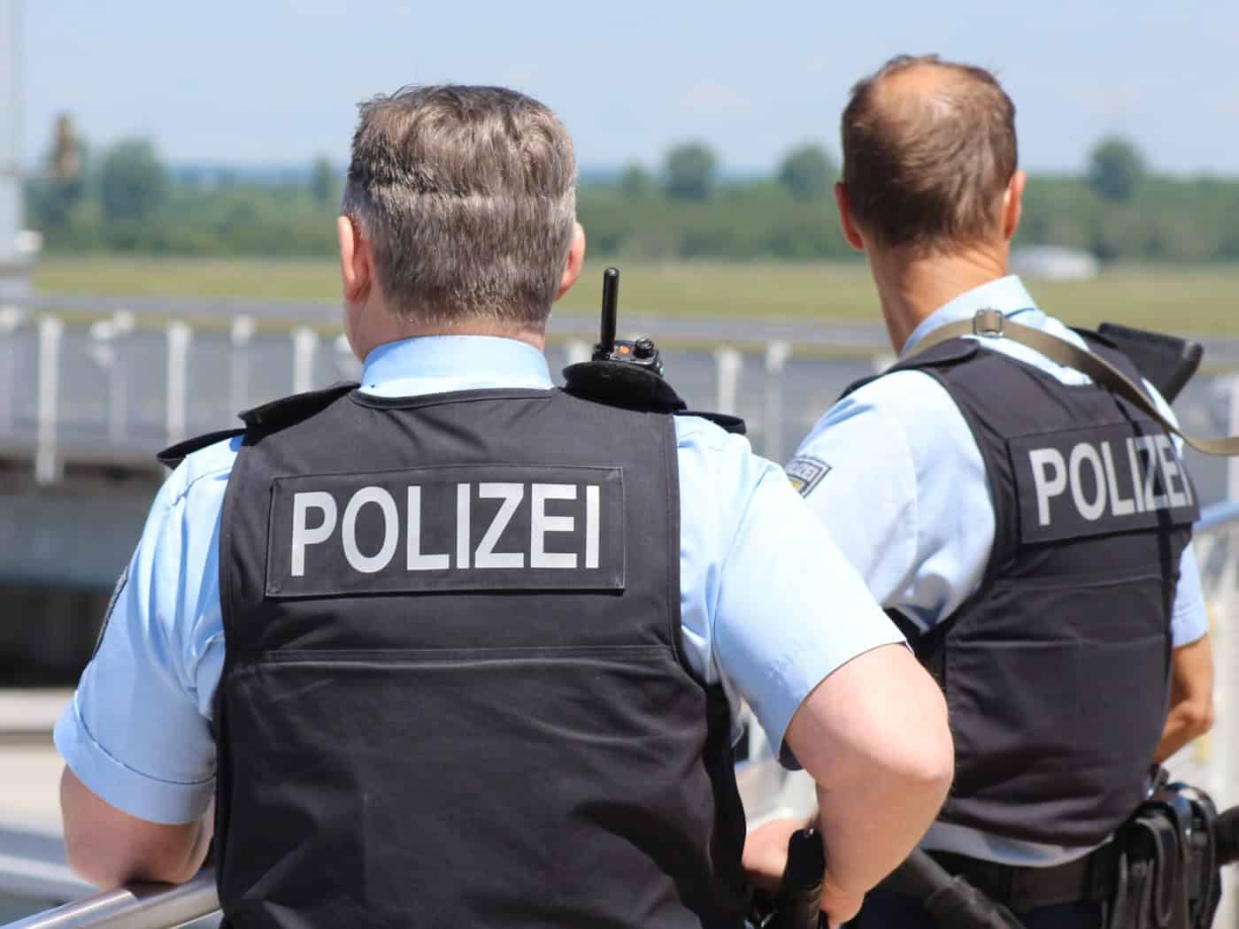 Rückenansicht von zwei Polizeibeamten in hellblauen Hemden und dunklen Schutzwesten mit der Aufschrift „POLIZEI“, die im Freien an einem Geländer stehen.