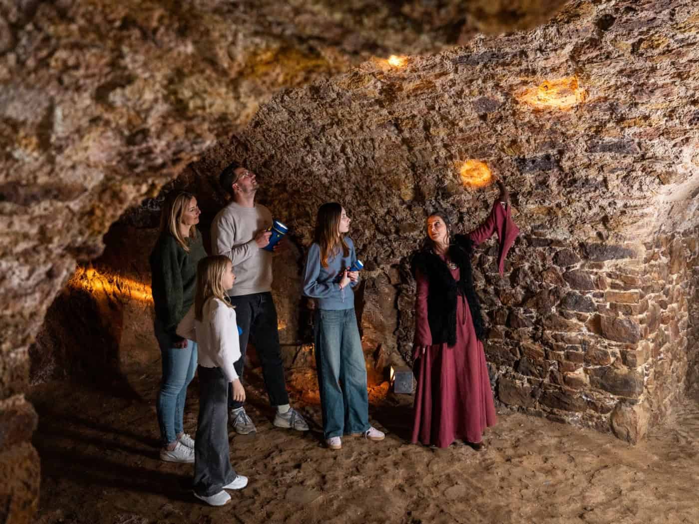 Eine Frau in historischem rotem Gewand zeigt einer Familie mit Taschenlampen das alte Kellergewölbe aus grobem Naturstein auf Burg Gnandstein. Die Decke wird von warmem Licht erhellt.
