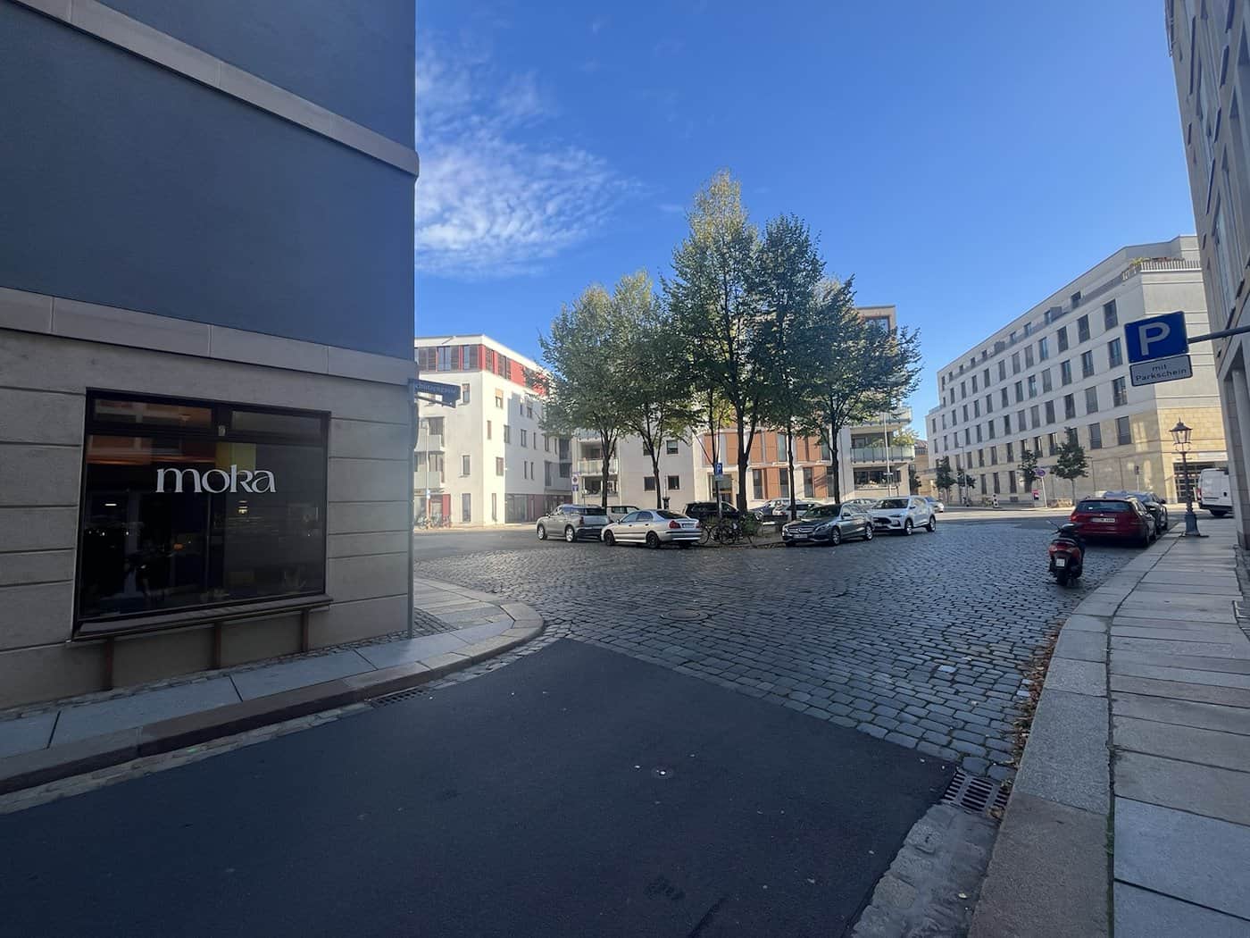 Blick in die Schützengasse in Dresden auf eine Straßenkreuzung und das Café Moka links im Bild; in der Mitte befindet sich eine von Autos umstellte, begrünte Verkehrsinsel (das grüne Dreieck) vor moderner Bebauung.