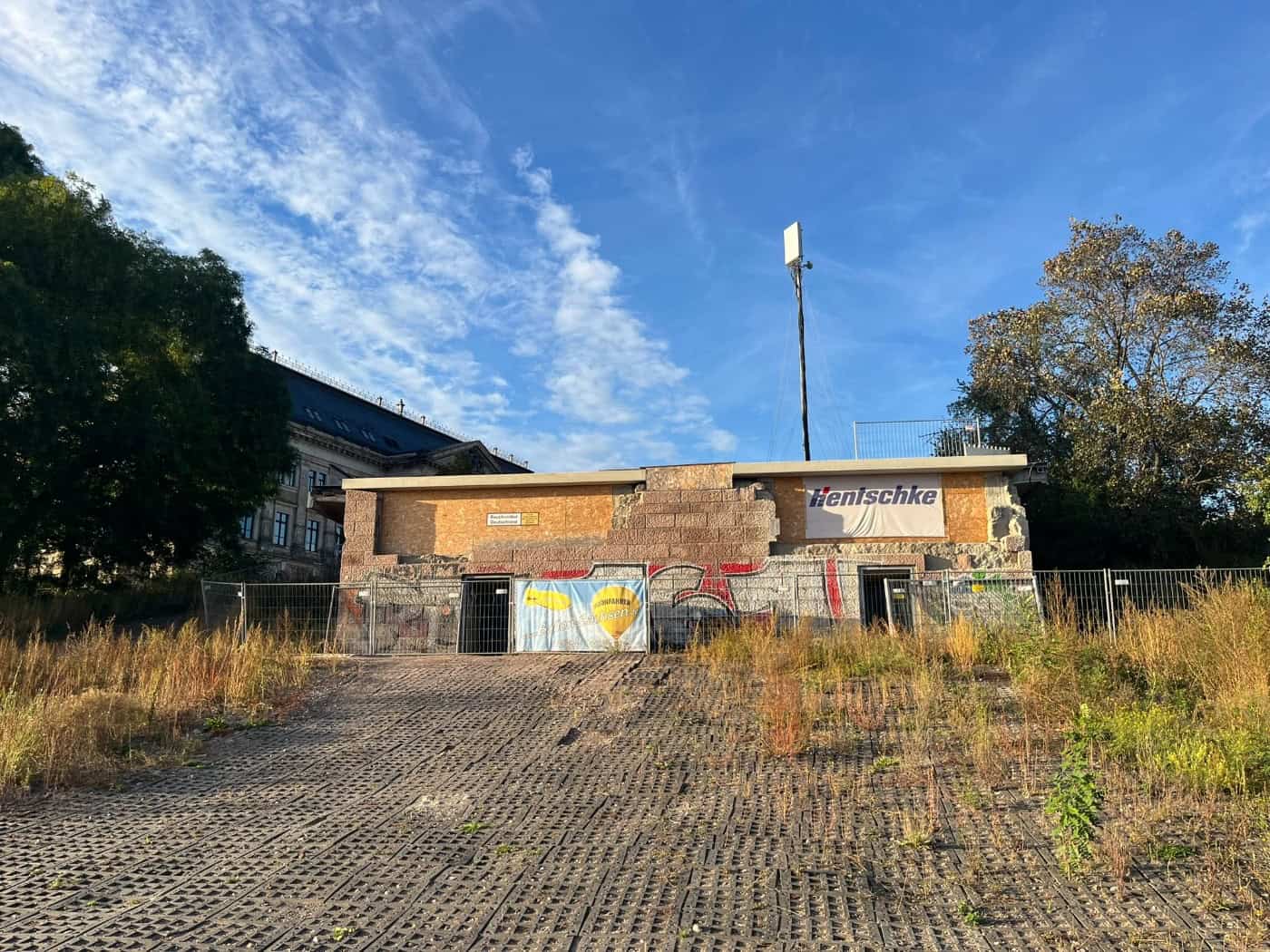 Abgesperrter Brückenkopf der eingestürzten Carolabrücke in Dresden mit Bauzäunen, Holzverkleidungen und Firmenbannern; davor befindet sich eine gepflasterte Rampe mit Wildwuchs unter blauem Himmel.