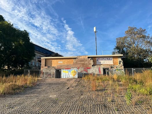 Abgesperrter Brückenkopf der eingestürzten Carolabrücke in Dresden mit Bauzäunen, Holzverkleidungen und Firmenbannern; davor befindet sich eine gepflasterte Rampe mit Wildwuchs unter blauem Himmel.
