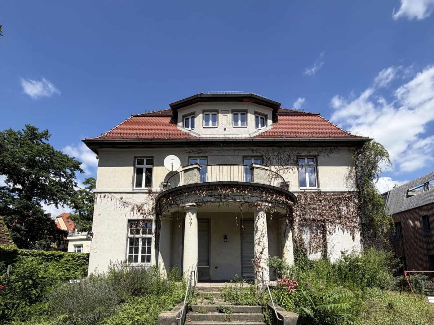 Vorderansicht einer zweigeschossigen, hellen Gründerzeit-Villa mit rotem Ziegeldach, Balkon mit Säulenportal im Eingangsbereich und leicht verwildertem Vorgarten unter blauem Himmel. (185 Zeichen)   SEO-Dateiname