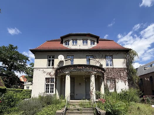 Vorderansicht einer zweigeschossigen, hellen Gründerzeit-Villa mit rotem Ziegeldach, Balkon mit Säulenportal im Eingangsbereich und leicht verwildertem Vorgarten unter blauem Himmel. (185 Zeichen)   SEO-Dateiname