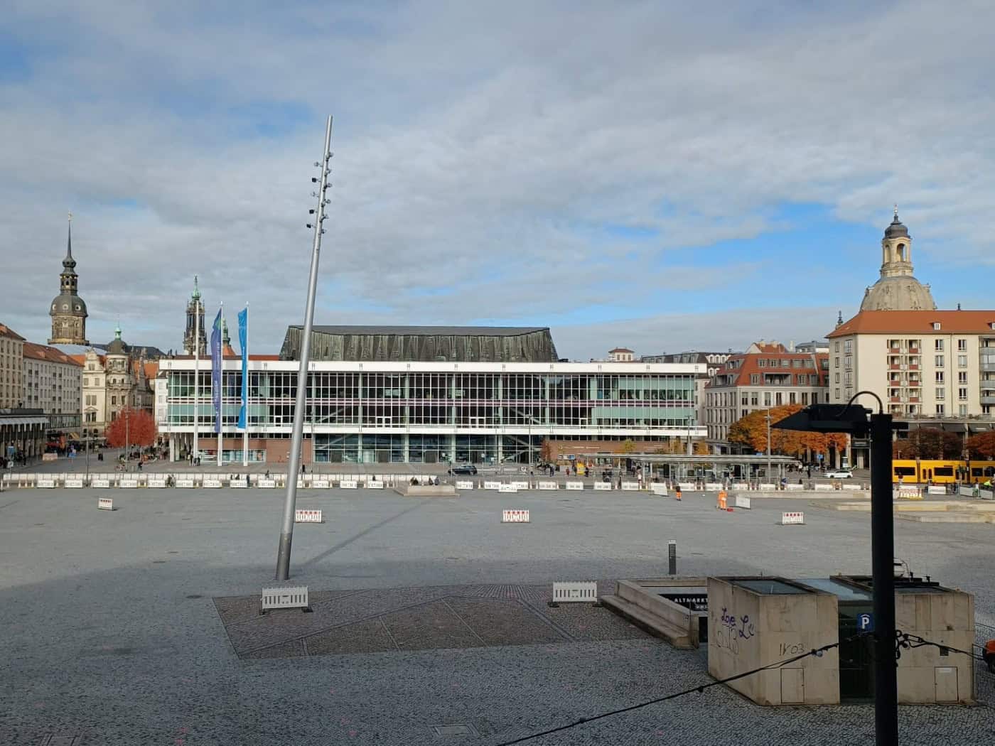 Weitwinkelblick über den weitgehend leeren, gepflasterten Dresdner Altmarkt bei Tageslicht. Im Hintergrund sind historische und moderne Gebäude wie der Kulturpalast sowie Kirchtürme der Altstadt unter einem blauen Himmel zu sehen.