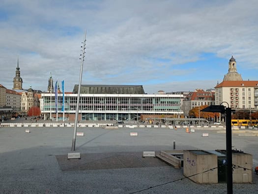 Weitwinkelblick über den weitgehend leeren, gepflasterten Dresdner Altmarkt bei Tageslicht. Im Hintergrund sind historische und moderne Gebäude wie der Kulturpalast sowie Kirchtürme der Altstadt unter einem blauen Himmel zu sehen.