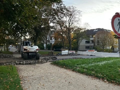 Ein weißer Baustellenbagger steht auf der aufgerissenen Fahrbahn der Mendelssohnallee in Dresden-Blasewitz. Umgeben von Grünflächen, Bäumen und Wohngebäuden wird der Abschnitt im Rahmen der Instandsetzung für den Verkehr gesperrt.