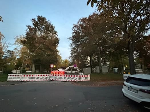 Rot-weiße Bauzäune und Verkehrsschilder sperren die Fahrbahn der Mendelssohnallee am Friedensplatz in Dresden-Blasewitz ab. Im Hintergrund stehen große Laubbäume, Baustellenfahrzeuge und ein weißes Auto rechts im Vordergrund.