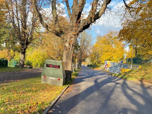 Ein grauer, mobiler Blitzeranhänger steht auf dem Grasstreifen unter herbstlichen Bäumen an der Blüherstraße in Dresden. Rechts verläuft die Straße am Rudolf-Harbig-Stadion vorbei, der Schatten des Fotografen zeichnet sich auf dem Asphalt ab.