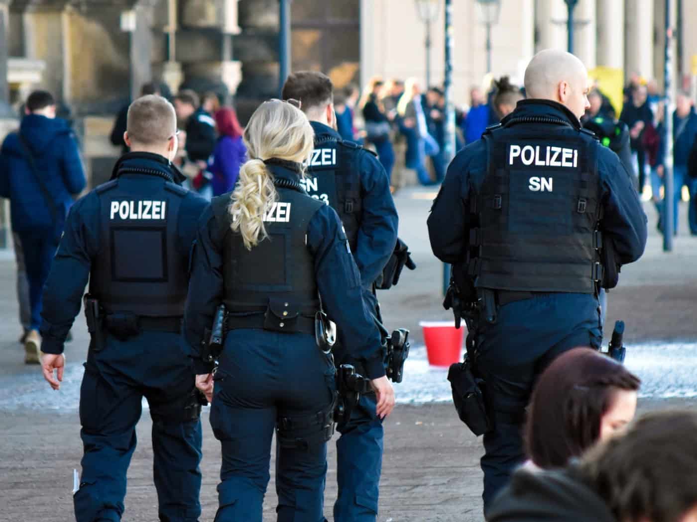 Symbolbild: Streife der Polizei in dunkelblauen Uniformen mit der Aufschrift „POLIZEI SN“ geht von hinten zu sehen durch eine belebte Fußgängerzone in Dresden. Im Hintergrund befinden sich Passanten und historische Sandsteingebäude bei Tageslicht.