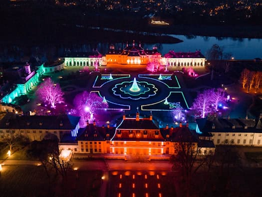Drohnenaufnahme von Schloss und Park Pillnitz bei Nacht: Der Lustgarten leuchtet in bunten Farben wie Magenta, Grün und Blau. In der Mitte steht ein hell erstrahlender Weihnachtsbaum, umgeben von den beleuchteten historischen Schlossgebäuden.