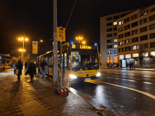 Nachtaufnahme bei nassem Asphalt: Ein gelber Gelenkbus der DVB-Linie 61 mit Fahrtziel Löbtau steht an einer beleuchteten Bushaltestelle. Im Vordergrund spiegelt sich Straßenlaternenlicht auf dem feuchten Pflaster und der Fahrbahn.