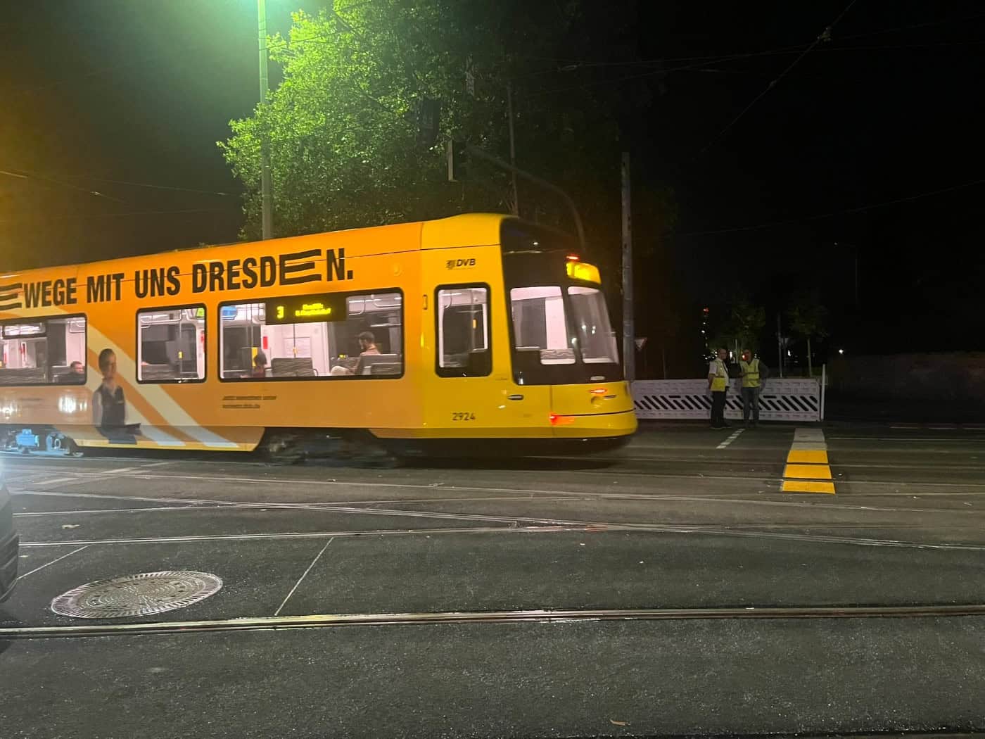 Eine gelbe DVB-Straßenbahn fährt bei Nacht über Gleise. Auf dem gelben Wagen prangt der Schriftzug „WEGE MIT UNS DRESDEN“. Im Hintergrund sind zwei Personen in fluoreszierenden Warnwesten an einer Straßenkreuzung zu sehen.