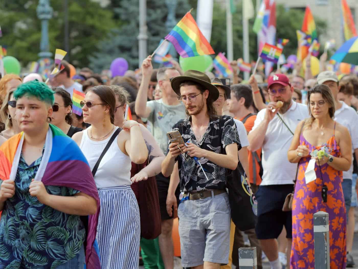Zahlreiche bunte Menschen ziehen bei einer Parade für die queere Community durch die Straße. Sie schwenken Regenbogenfahnen und tragen Regenbogen-Accessoires, darunter ein junger Mann mit grünem Haar und ein Mann mit Hut und Brille.