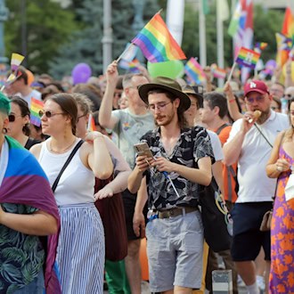 Zahlreiche bunte Menschen ziehen bei einer Parade für die queere Community durch die Straße. Sie schwenken Regenbogenfahnen und tragen Regenbogen-Accessoires, darunter ein junger Mann mit grünem Haar und ein Mann mit Hut und Brille.