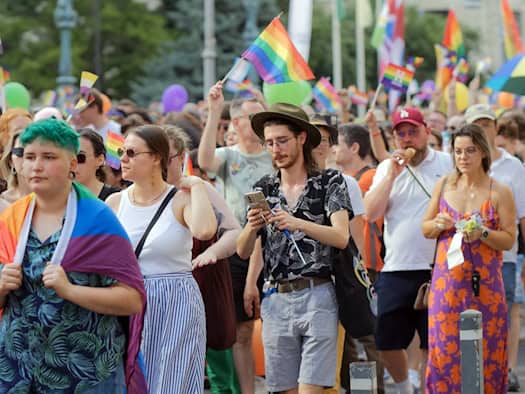 Zahlreiche bunte Menschen ziehen bei einer Parade für die queere Community durch die Straße. Sie schwenken Regenbogenfahnen und tragen Regenbogen-Accessoires, darunter ein junger Mann mit grünem Haar und ein Mann mit Hut und Brille.