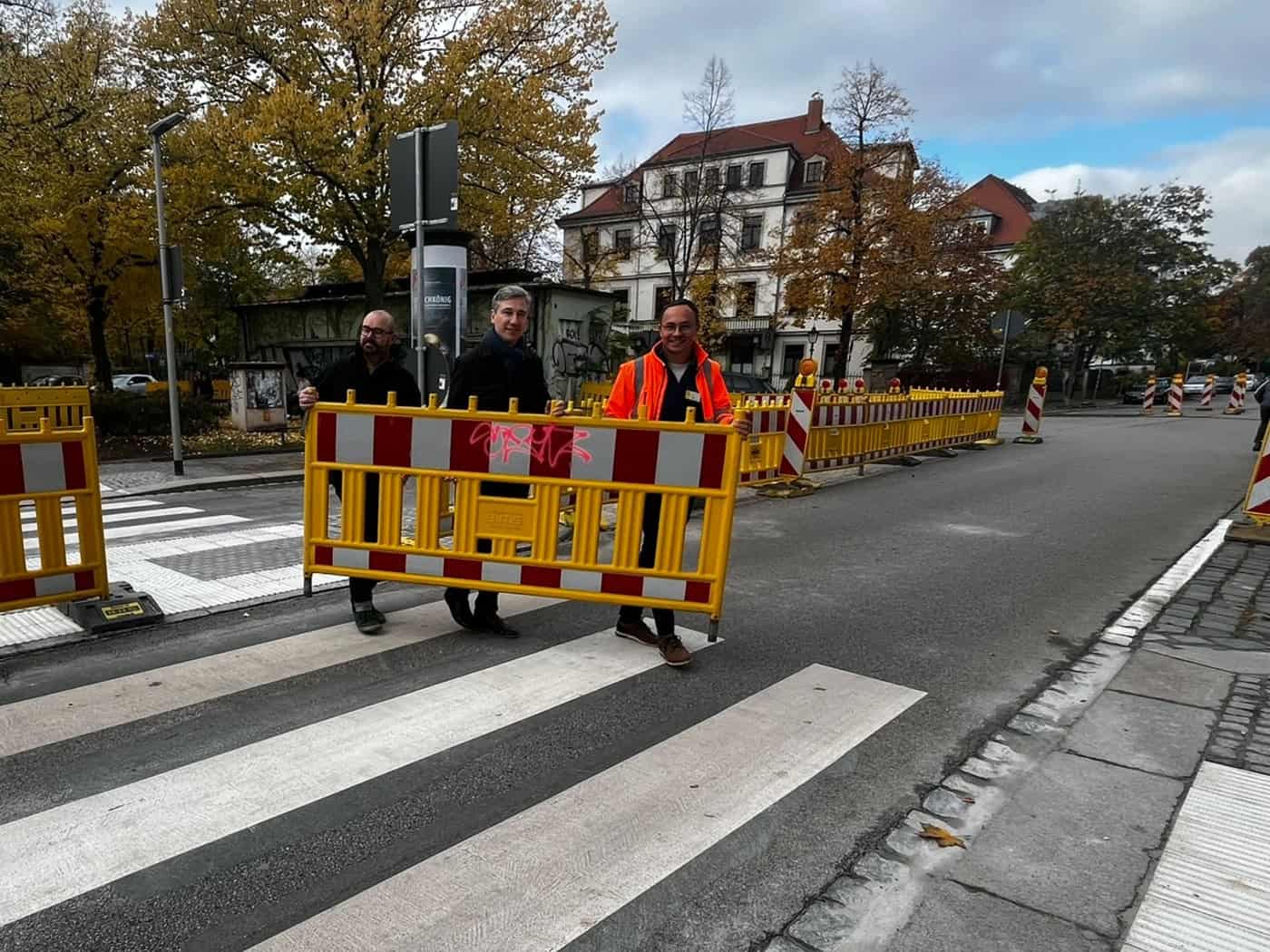 Drei Männer tragen gemeinsam eine gelb-rote Baustellenbarke von einer frisch markierten Zebrastreifen-Insel auf der Straße. V.l.n.r.: Stefan Kraft, Stephan Kühn und Ralf Langer in Warnweste vor herbstlicher Kulisse am Heinrich-Schütz-Konservatorium.