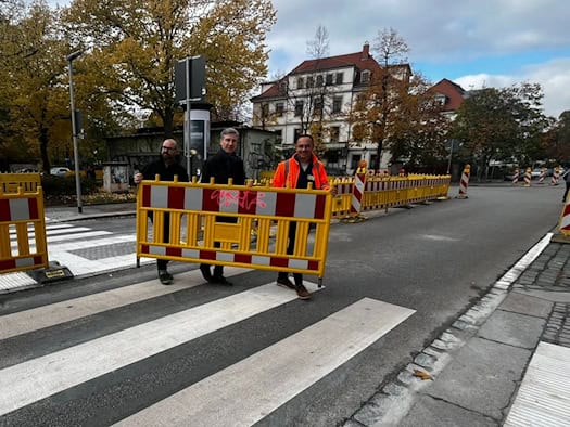 Drei Männer tragen gemeinsam eine gelb-rote Baustellenbarke von einer frisch markierten Zebrastreifen-Insel auf der Straße. V.l.n.r.: Stefan Kraft, Stephan Kühn und Ralf Langer in Warnweste vor herbstlicher Kulisse am Heinrich-Schütz-Konservatorium.