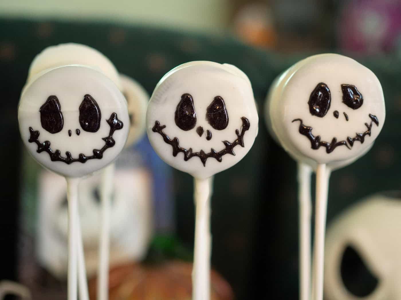 Drei weiß glasierte Cake-Pops am Stiel in einer Reihe als Grusel-Süßigkeit. Auf der Glasur sind mit dunkler Schokolade grinsende Skelett-Gesichter im Totenkopf-Stil aufgemalt. Der Hintergrund zeigt unscharfe Halloween-Dekorationen.