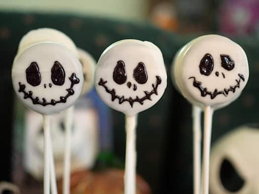 Drei weiß glasierte Cake-Pops am Stiel in einer Reihe als Grusel-Süßigkeit. Auf der Glasur sind mit dunkler Schokolade grinsende Skelett-Gesichter im Totenkopf-Stil aufgemalt. Der Hintergrund zeigt unscharfe Halloween-Dekorationen.