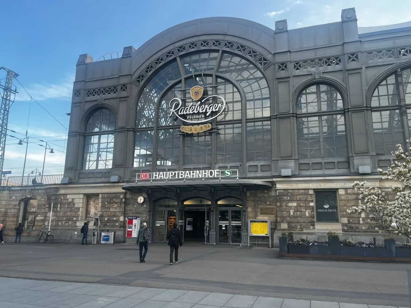 Außenansicht der historischen Glas- und Sandsteinfassade des Dresdner Hauptbahnhofs mit dem Schriftzug „HAUPTBAHNHOF” und großer Werbeaufschrift. Personen gehen vor den Eingangstüren des Bahnhofsgebäudes auf dem Bahnhofsvorplatz