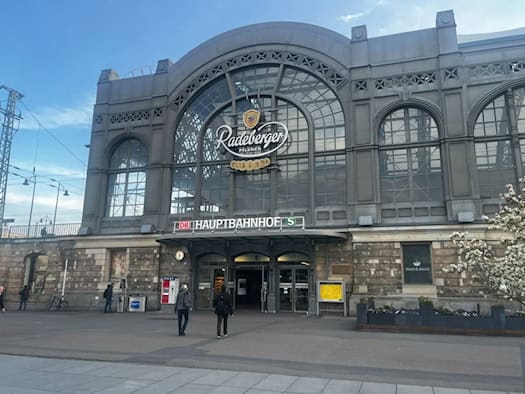 Außenansicht der historischen Glas- und Sandsteinfassade des Dresdner Hauptbahnhofs mit dem Schriftzug „HAUPTBAHNHOF” und großer Werbeaufschrift. Personen gehen vor den Eingangstüren des Bahnhofsgebäudes auf dem Bahnhofsvorplatz