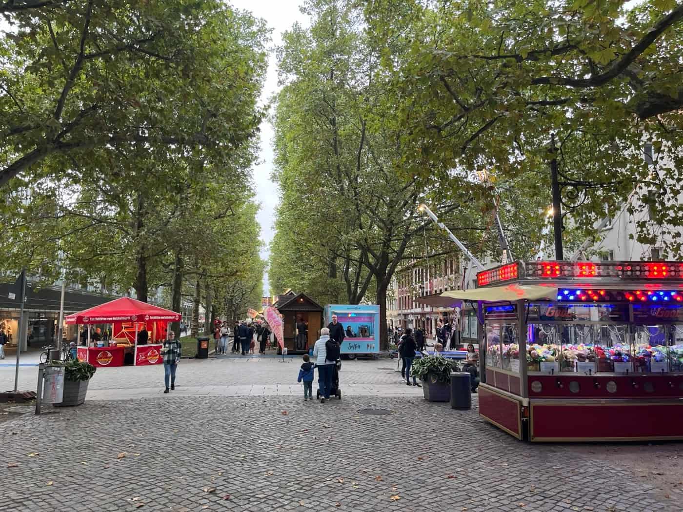 Blick über die von hohen Bäumen gesäumte Dresdner Hauptstraße während des Familienfestes. Auf dem Pflaster stehen bunte Verkaufsstände, Imbissbuden und ein beleuchteter Schaustellerwagen. Einige Passanten flanieren entlang der Allee.