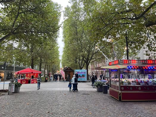 Blick über die von hohen Bäumen gesäumte Dresdner Hauptstraße während des Familienfestes. Auf dem Pflaster stehen bunte Verkaufsstände, Imbissbuden und ein beleuchteter Schaustellerwagen. Einige Passanten flanieren entlang der Allee.