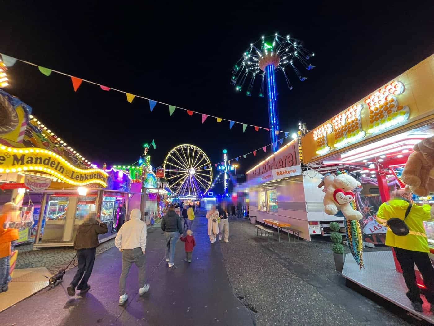 Abendliche Ansicht des Herbstvolksfests an der Pieschener Allee in Dresden bei Dunkelheit. Bunt beleuchtete Buden, Spielgeschäfte und Verkaufsstände säumen den Weg, im Hintergrund leuchtet das große Riesenrad am Nachthimmel.
