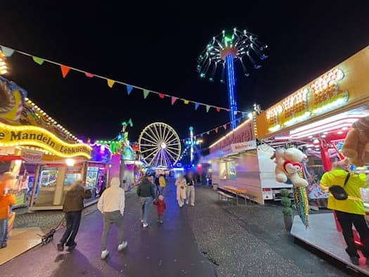 Abendliche Ansicht des Herbstvolksfests an der Pieschener Allee in Dresden bei Dunkelheit. Bunt beleuchtete Buden, Spielgeschäfte und Verkaufsstände säumen den Weg, im Hintergrund leuchtet das große Riesenrad am Nachthimmel.