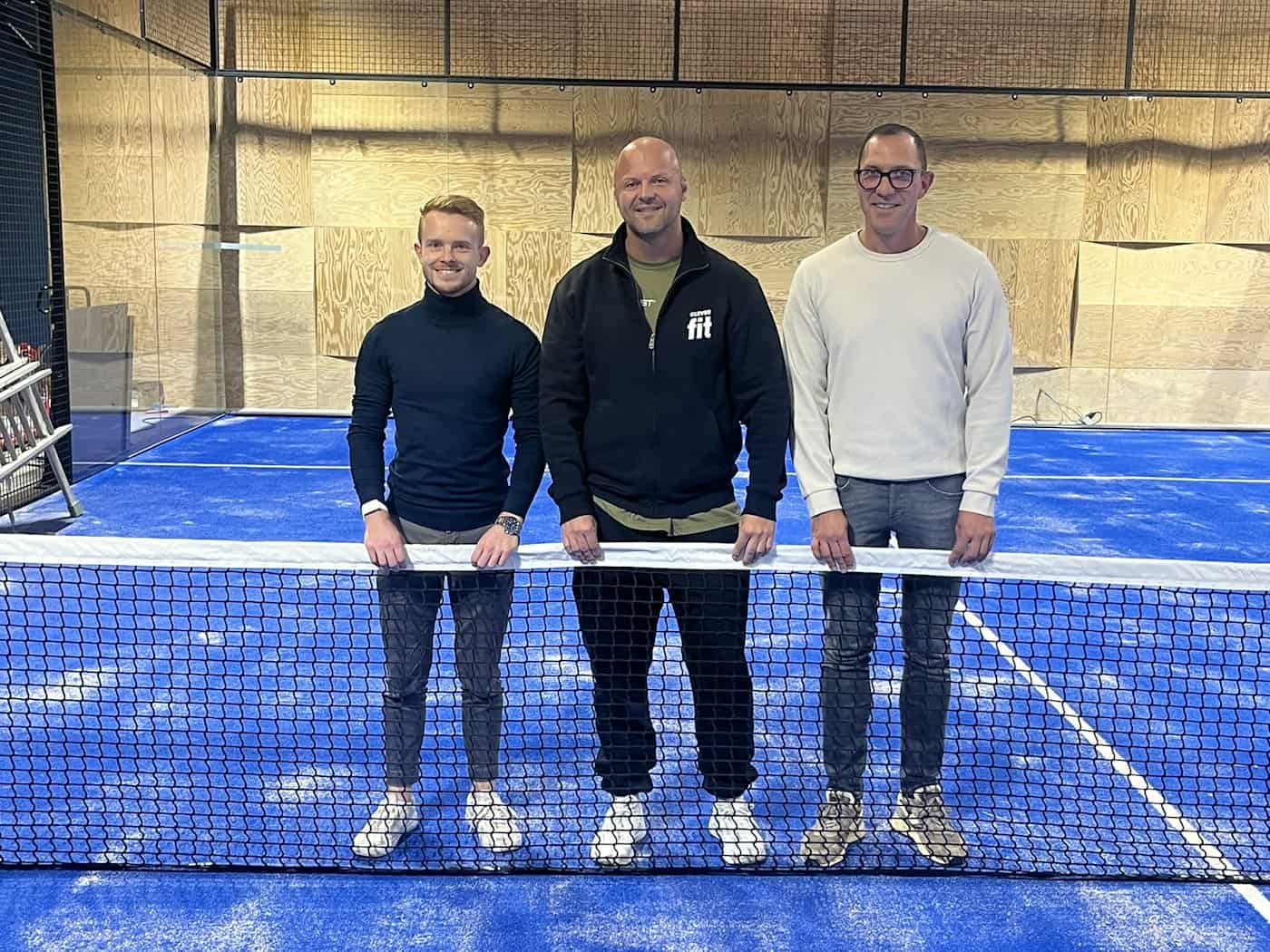 Drei Männer stehen lächelnd hinter dem Netz eines blauen Indoor-Padel-Courts im ehemaligen Bofimax-Kino Dresden. Im Hintergrund sind Glaswände und Holzpaneele der Halle zu sehen.
