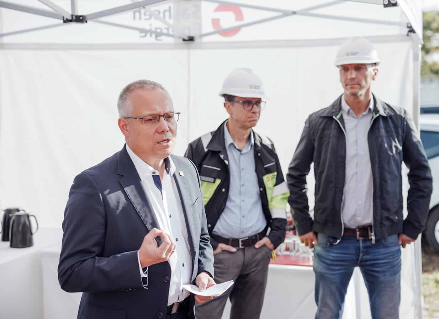 Wirtschaftsbürgermeister Jan Pratzka informiert sich zum Stromnetzausbau in Dresden Dresdens Wirtschaftsbürgermeister Jan Pratzka spricht im dunklen Sakko mit Notizen in der Hand unter einem weißen Pavillon. Hinter ihm stehen zwei Männer mit weißen Schutzhelmen.