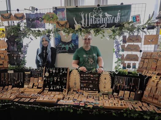 Zwei Ausstellerinnen (v.l.n.r. eine Frau mit blauen Haaren und eine Frau in grünem Oberteil) stehen hinter einem reich dekorierten Stand. Auf Tischen und Aufstellern präsentieren sie handgefertigten Schmuck, Ketten und Kunsthandwerk vor einem Banner.