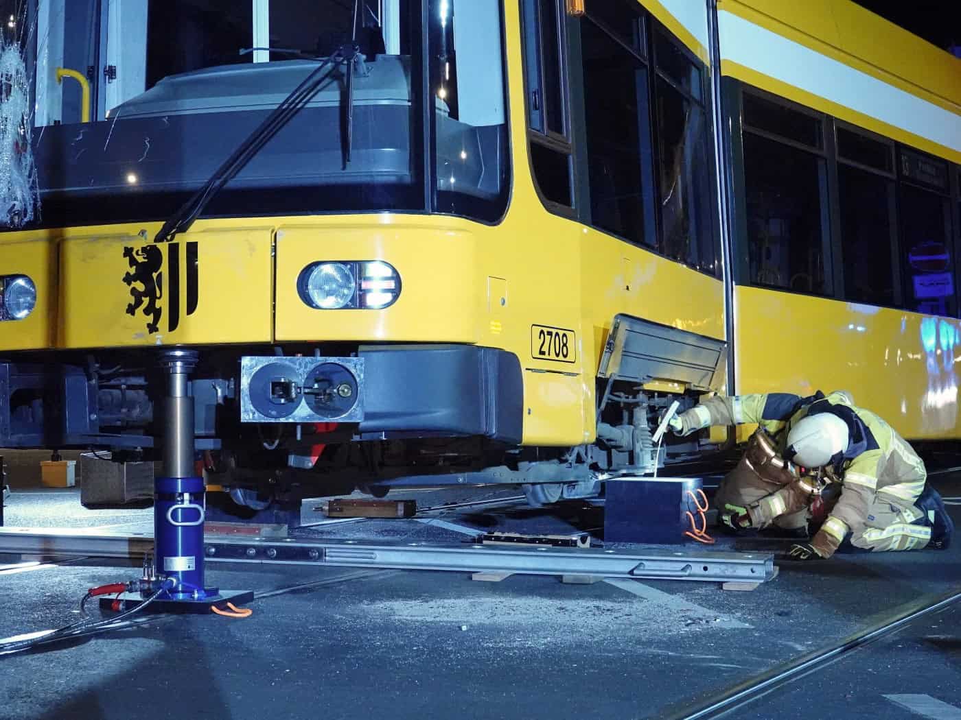 Nachtaufnahme einer beschädigten gelben DVB-Straßenbahn in Dresden, die mit blauer Hydraulik-Hebetechnik angehoben wird. Ein Feuerwehrmann in Schutzkleidung kniet am Boden und kontrolliert das Fahrwerk an den Schienen auf der gesperrten Kreuzung.