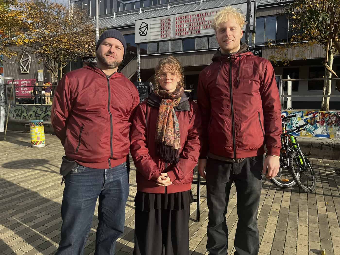 Kiezlichter-Koordinator Alessandro Finke (links) steht gemeinsam mit zwei weiteren Teammitgliedern in roter Projektkleidung auf einem gepflasterten Platz im Freien. Im Hintergrund ist ein Veranstaltungsplakat in der Dresdner Neustadt zu sehen.