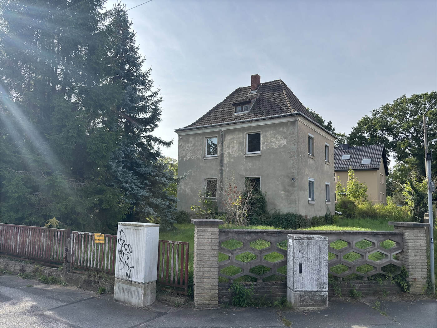 Einfamilienhaus Waldteichstraße 8 in Dresden-Klotzsche: Verkauf ab 200.000 Euro Unsaniertes, zweigeschossiges Einfamilienhaus mit schlichtem Grauputz und Walmdach aus dem Jahr 1930. Das Gebäude steht auf einem eingewachsenen Grundstück hinter einem Beton- und Holz-Gartenzaun unter leichtem Sonnenlicht.